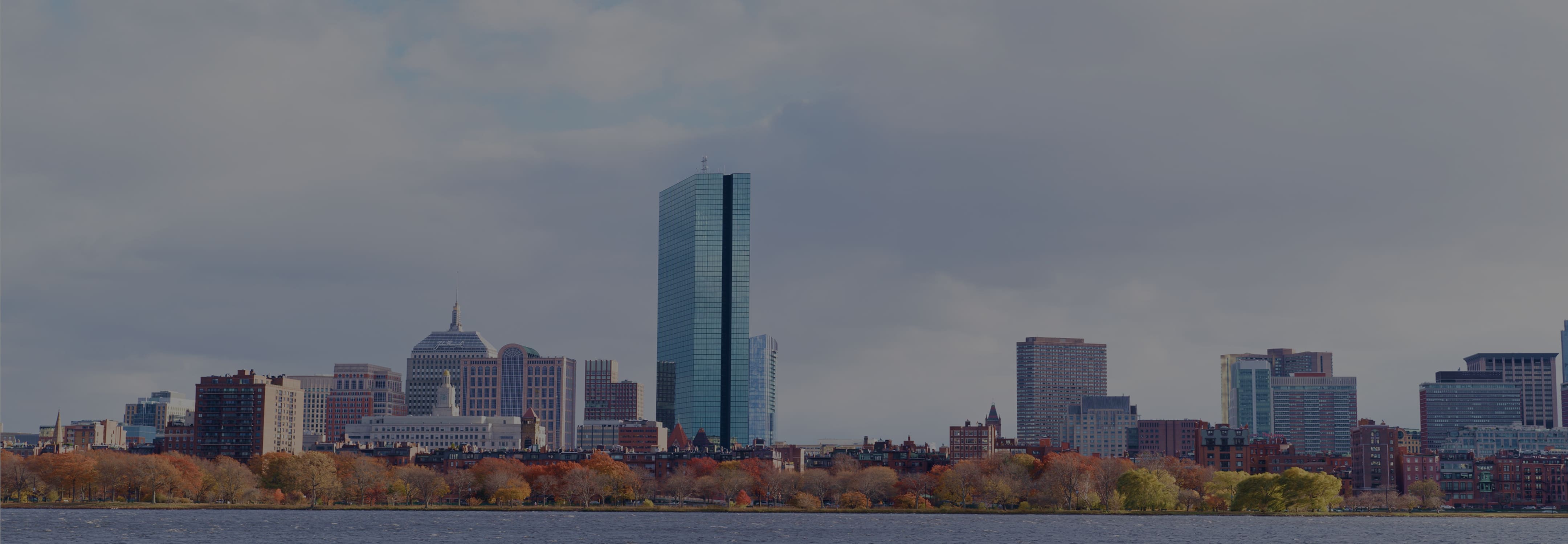 Boston skyline across the Charles River on a cloudy day, with autumn trees along the waterfront.