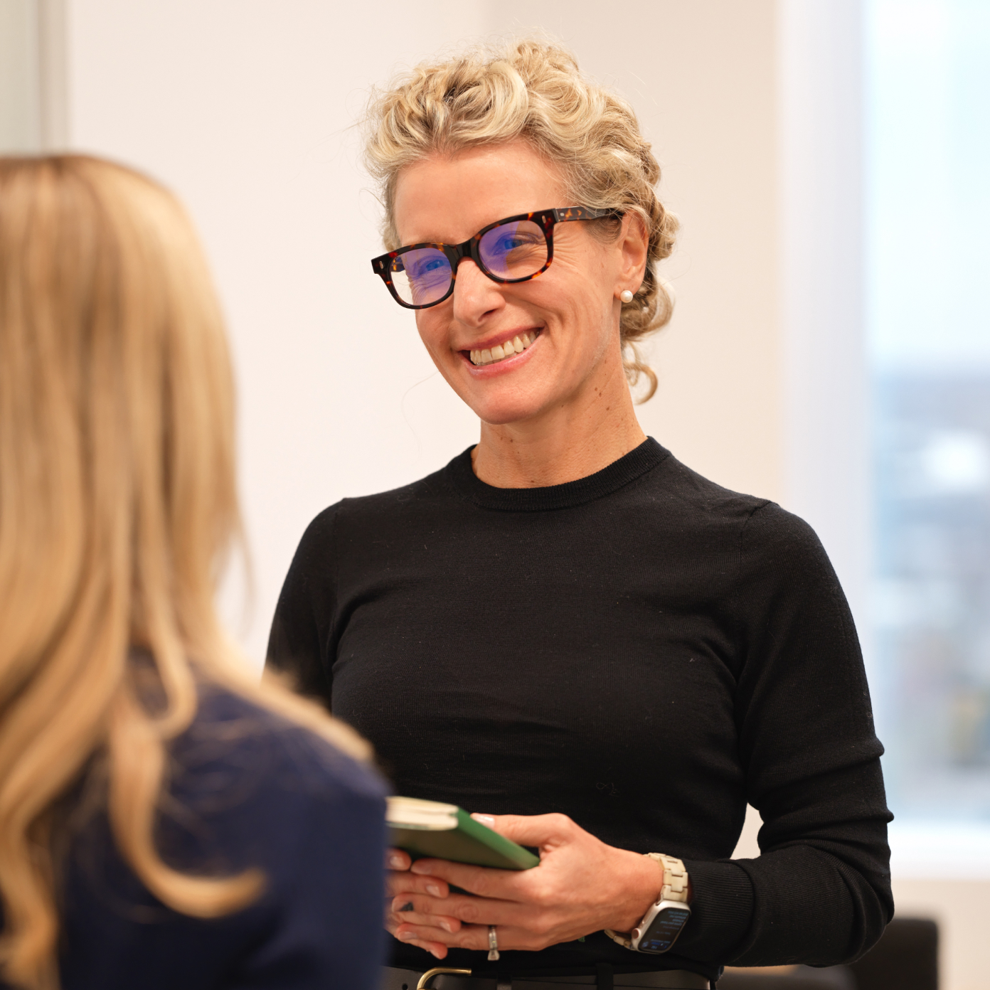 Smiling colleague with glasses holding a notebook while talking in a modern office.