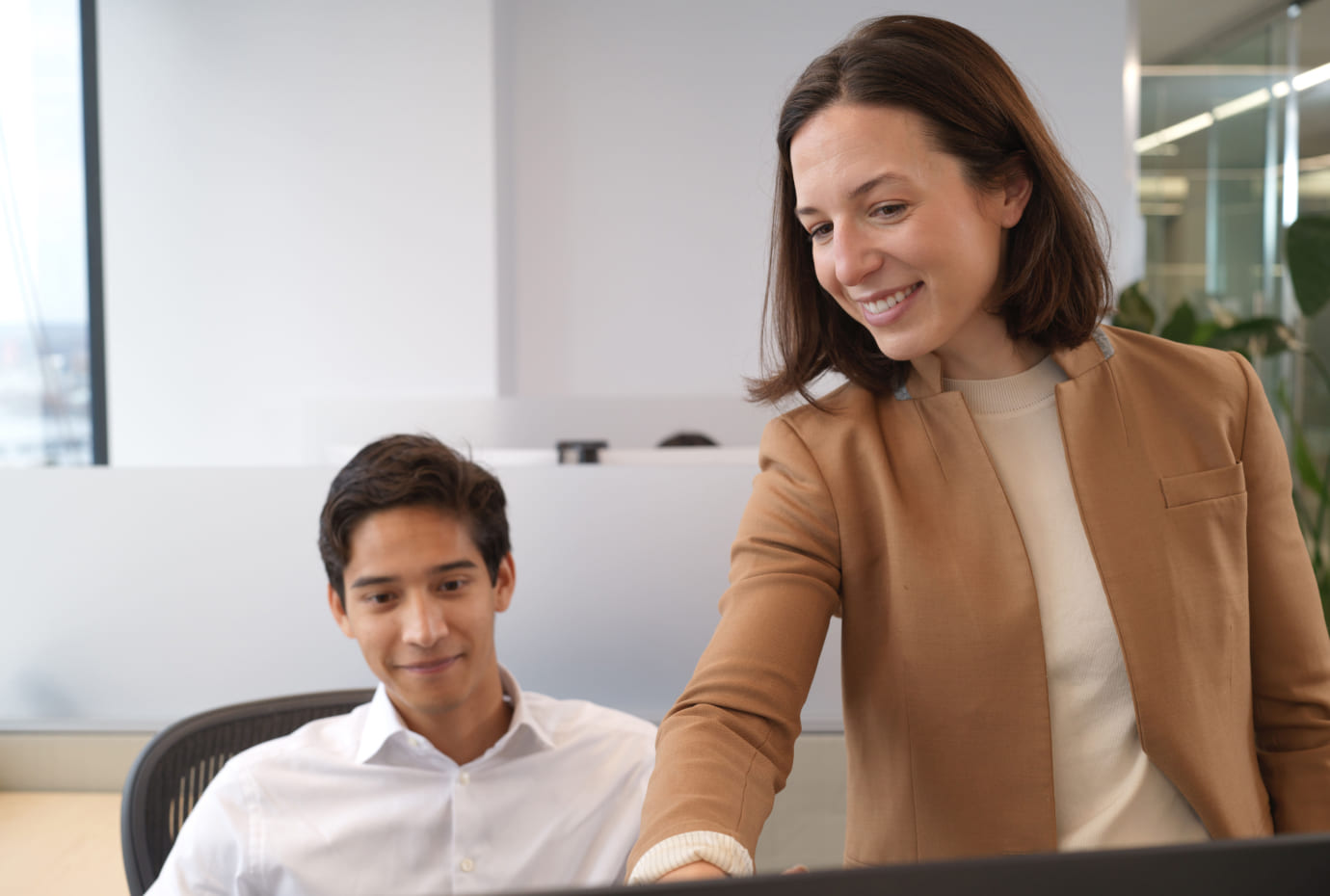 A colleague shares feedback at a desk while a teammate looks on in a bright office.