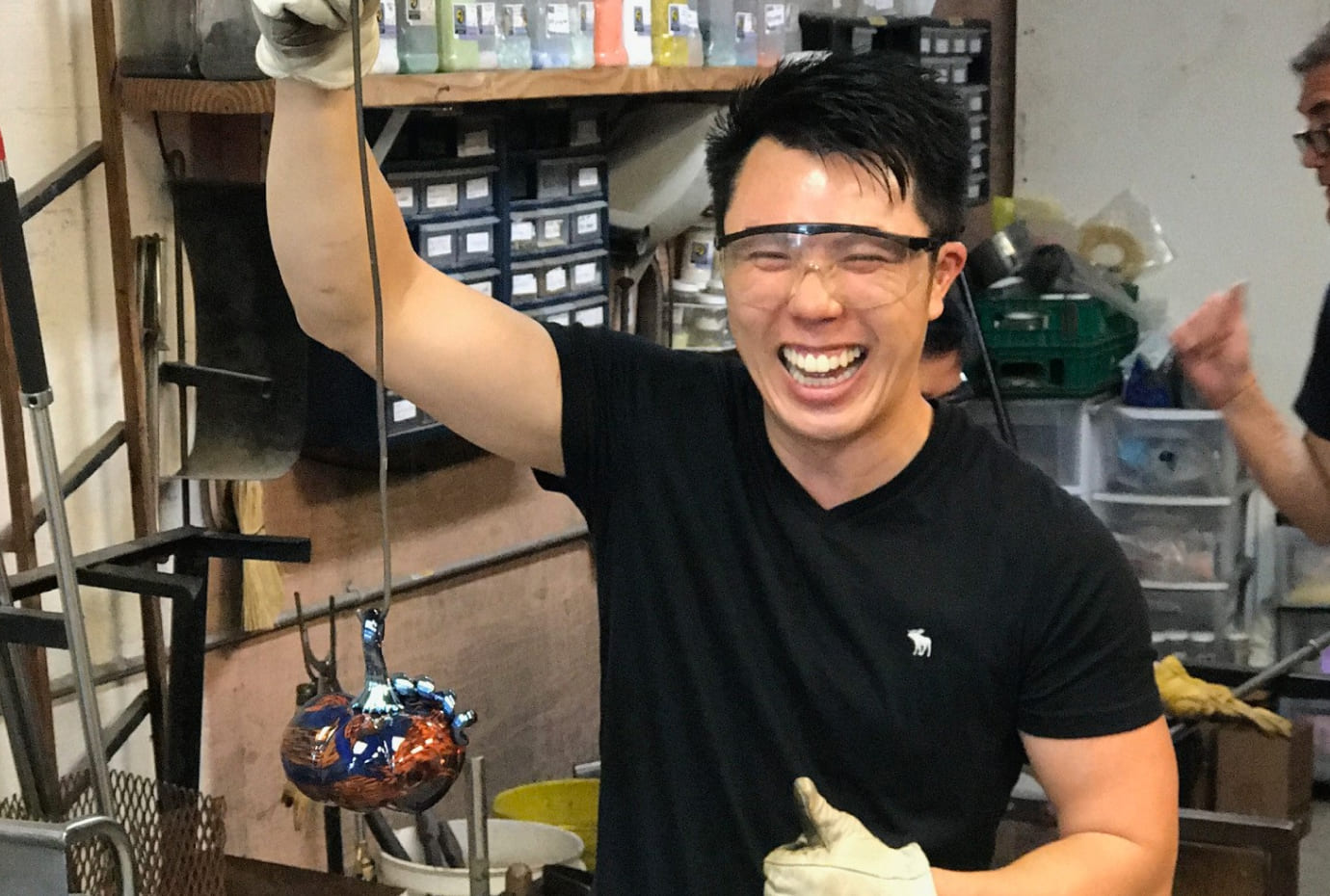 Person in a workshop lifts a multicolored molten glass piece with a rod, wearing protective eyewear.