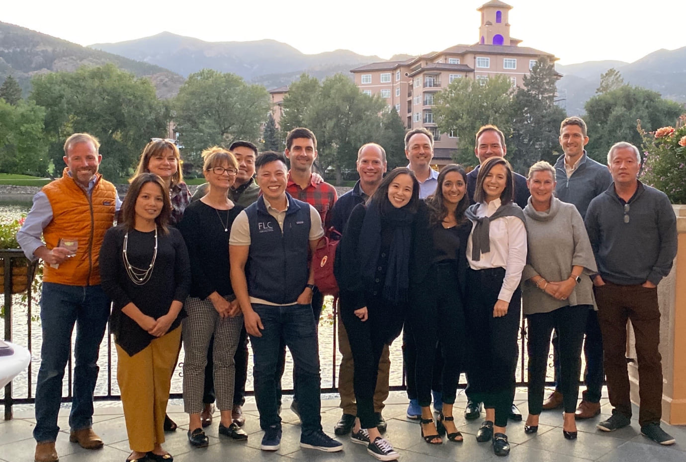 Group photo of colleagues outdoors by a lake with mountains and a resort building in the background.