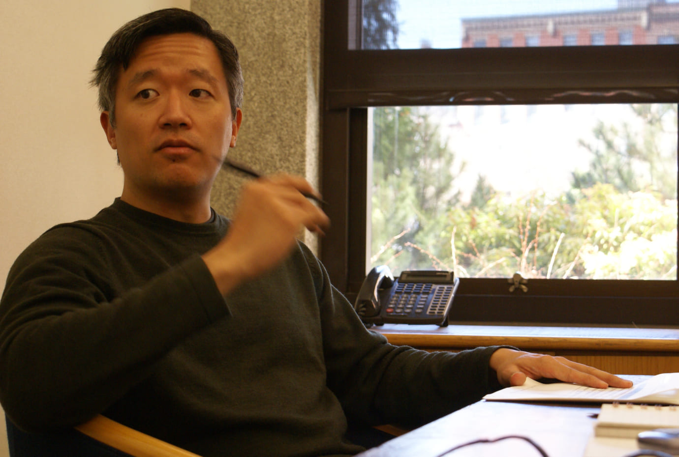 Colleague holding a pen during a meeting at a desk, with a window view in the background.