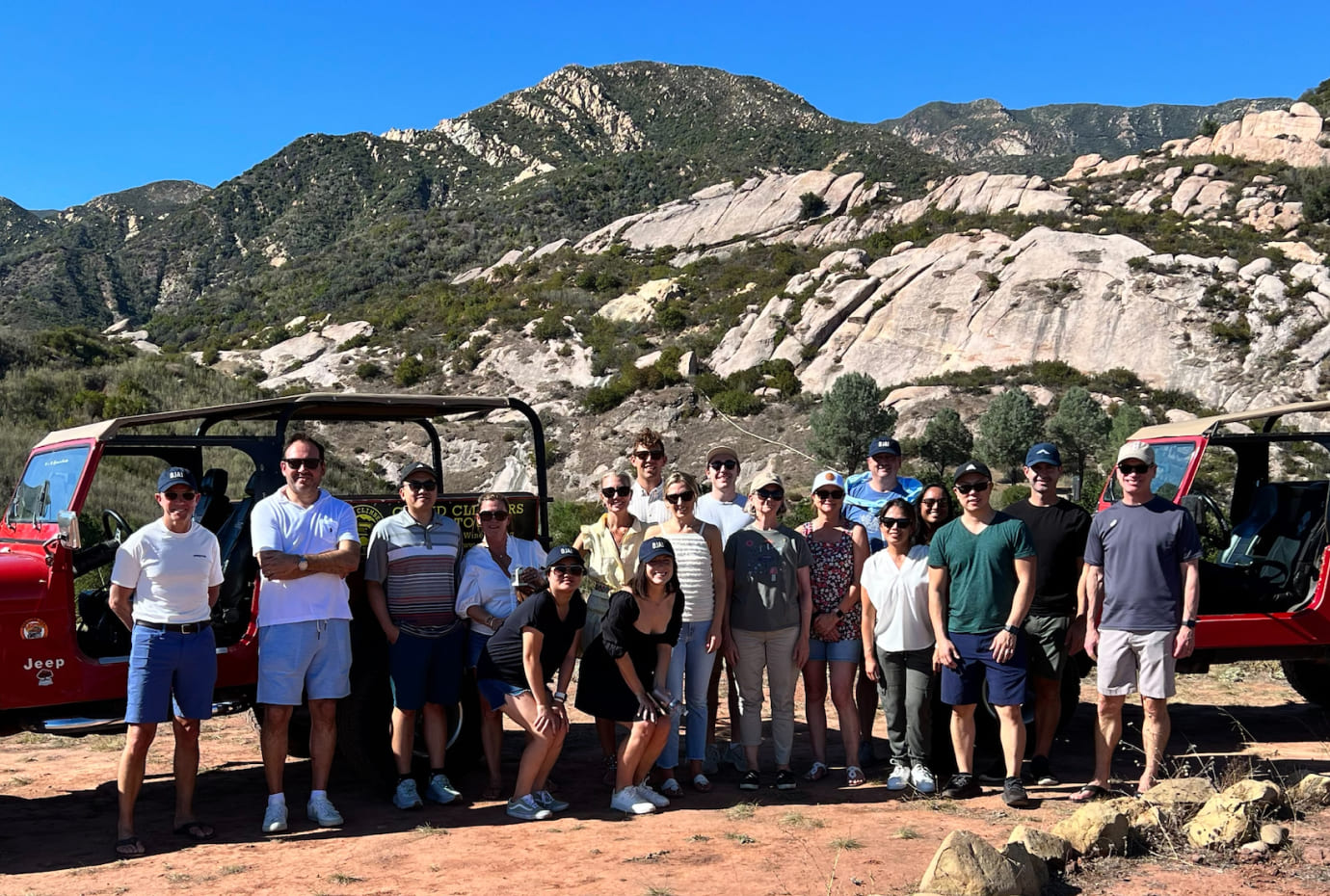 Group photo of colleagues on an outdoor off-road trip, standing between red Jeeps with rocky mountains in the background.