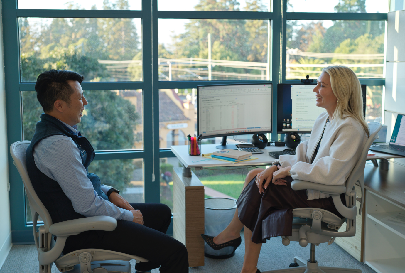 Two colleagues having a conversation in an office, seated at a desk with dual monitors and large windows.